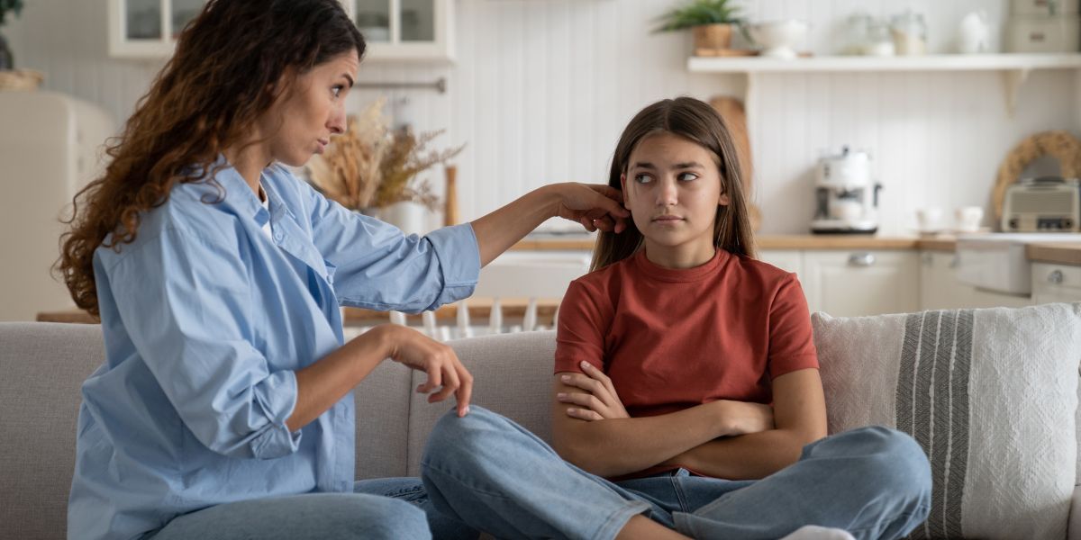 Mother pointing and speaking firmly to her teenage daughter, who sits with arms crossed and an irritated expression.